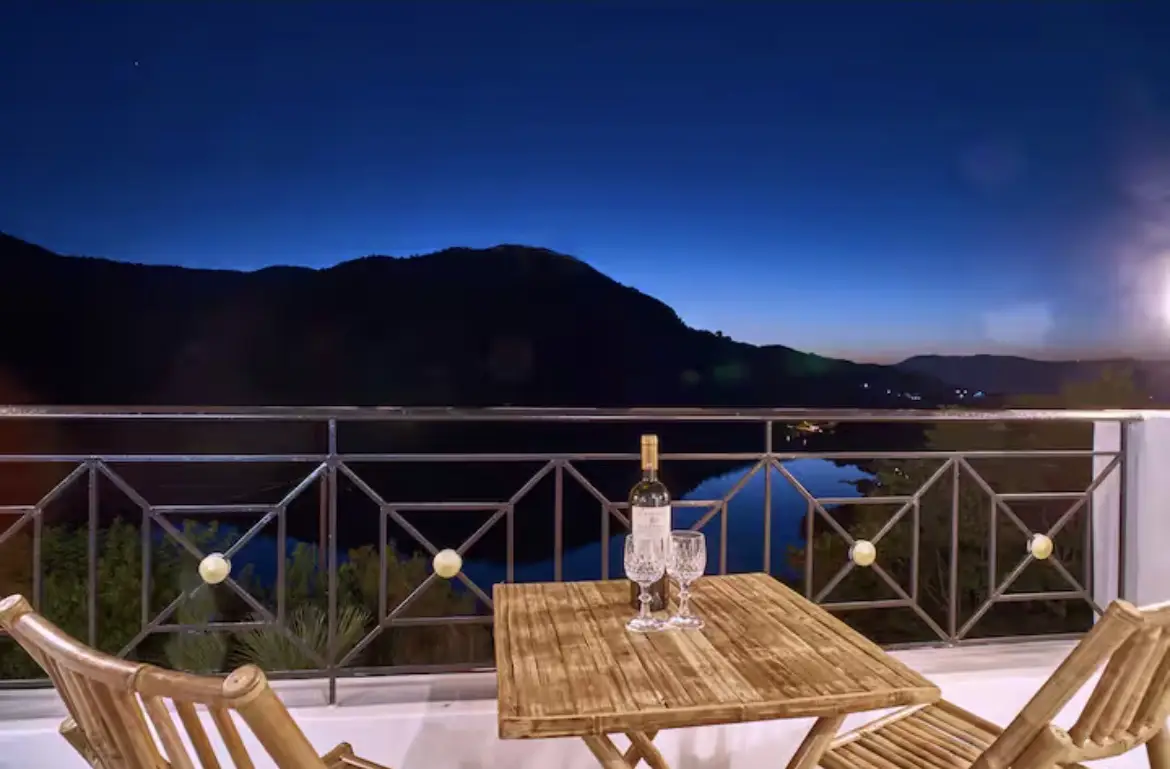 A wooden table with wine glasses and a bottle of wine on a private balcony overlooking the illuminated Lake Kournas at dusk.