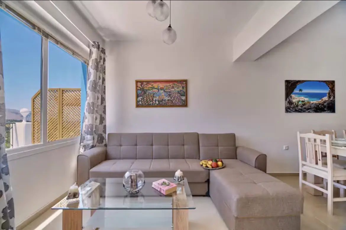 Modern living room in Suite 2 featuring a large L-shaped beige fabric sofa, glass coffee table, dining area, and bright window views.