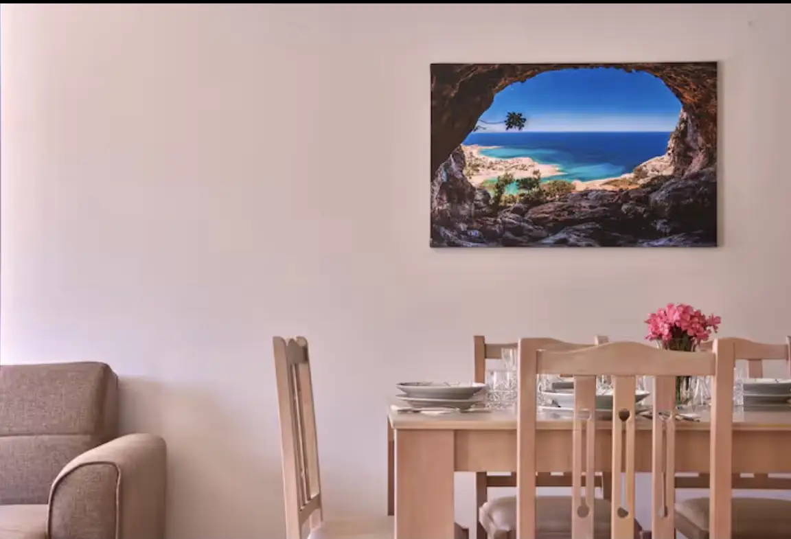 A minimalist dining setup in Suite 2 with light wood chairs and table, featuring a beautiful picture of a blue ocean cove on the wall.